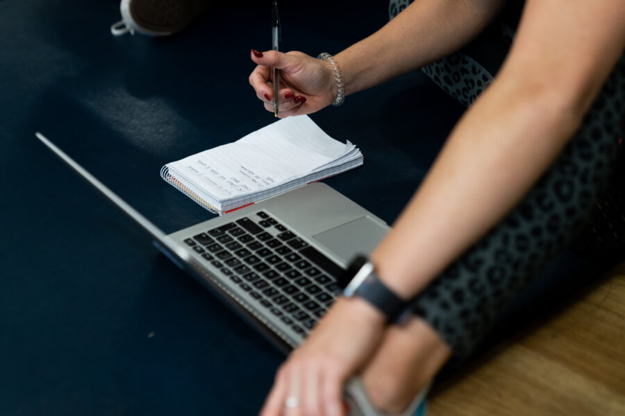 Close up of women's arms learning over her leg wearing active wear leggings, writing down exercises onto a pad of lined paper from an open laptop.