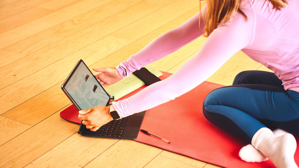 Young woman wearing a pink tight fitting fitness top sitting on a yoga mat adjusting a tablet screen, showing exercises