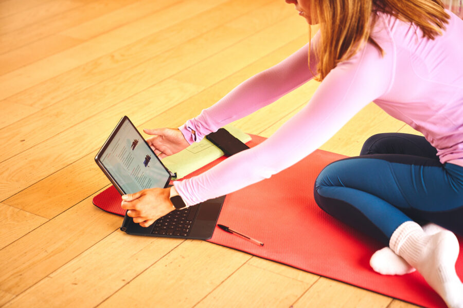 Young woman wearing a pink tight fitting fitness top sitting on a yoga mat adjusting a tablet screen, showing exercises