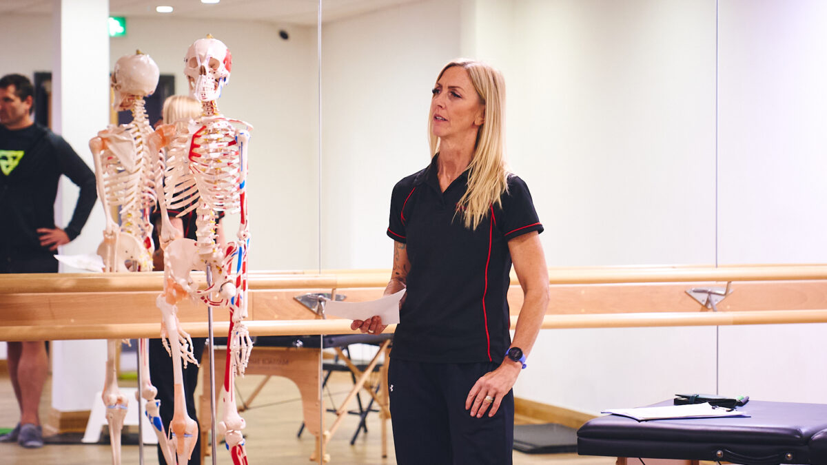 A fitness instructor with long blonde hair is standing in a gym, holding a piece of paper and speaking. Beside her is a human skeleton model.