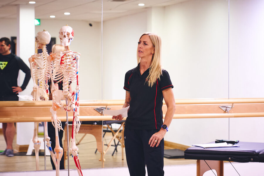 A fitness instructor with long blonde hair is standing in a gym, holding a piece of paper and speaking. Beside her is a human skeleton model.