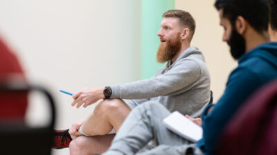 man with red hair and beard, sat down in a gym environment, wearing a grey hoody, and holding a pen, he is engaging in a talk with someone off camera.